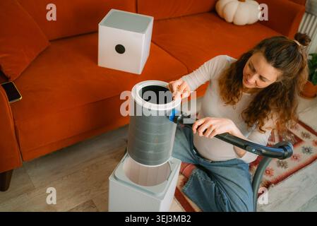 Woman cleaning an air purifier filter with a vacuum cleaner and brush attachment, performing routine maintenance to improve indoor air quality and ensure clean breathing in a modern room Stock Photo