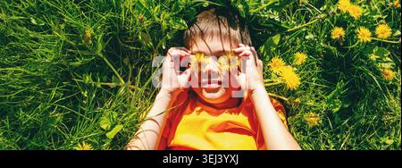 Funny boy lying on green grass and covering his eyes with yellow dandelions. Stock Photo