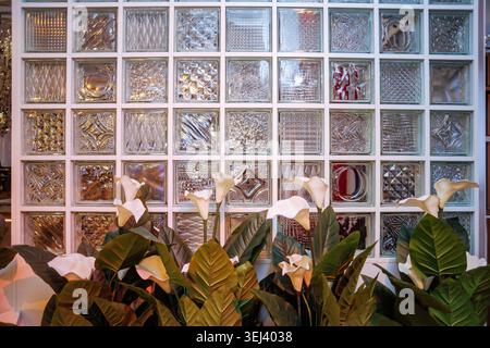 A front-facing shot of artificial white calla lily flowers arranged before a modern wall made of textured glass bricks Stock Photo