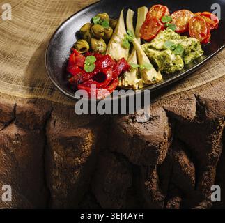 Food, Fresh vegetables including artichokes, olives, roasted peppers, and tomatoes are beautifully arranged on a black plate atop a rustic wooden surf Stock Photo