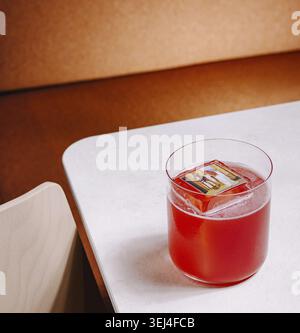 Food, A vibrant red cocktail with a square ice cube resting on top is placed on a white table. The warm backdrop creates an inviting atmosphere during Stock Photo