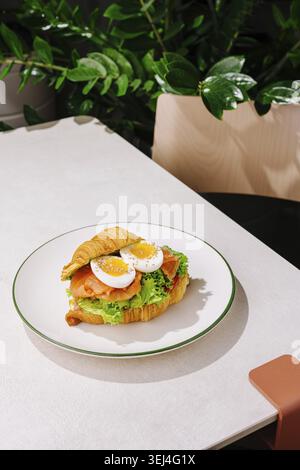 Food, A croissant sandwich filled with lettuce, tomato, and sliced boiled eggs sits on a simple white plate. The table features a plant backdrop, addi Stock Photo