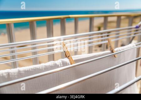 White laundry hanging on drying rack with clothespins on sunny balcony overlooking beach and turquoise sea in summer Stock Photo