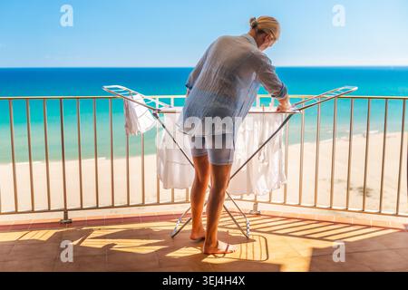 Woman hanging freshly washed clothes on drying rack on sunny balcony with sea view during summer vacation at beach apartment Stock Photo