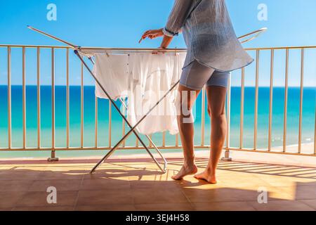 Woman hanging white laundry on drying rack on sunny balcony with turquoise sea view during summer vacation by the beach Stock Photo