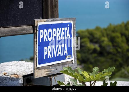 Vieste - Apulia - Italy - 08-29-2025: Blue sign reading PROPRIETA PRIVATA is fixed to weathered wood, with turquoise sea and green trees softly blurre Stock Photo