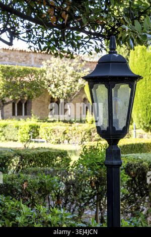 Vieste - Apulia - Italy - 08-29-2025: Black metal garden lantern stands beneath leafy branches, with trimmed hedges and a stone building with arched w Stock Photo