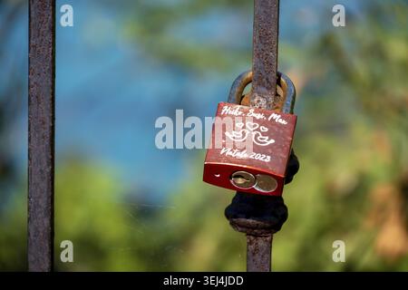 Vieste - Apulia - Italy - 08-29-2025: Red padlock with handwritten names and hearts hangs on a rusty iron fence, with blurred green and blue backgroun Stock Photo