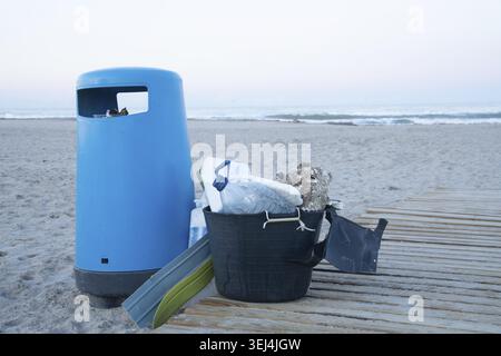 A blue waste bin next to a black plastic container with trash including styrofoam and plastic bottles on a wooden dock at the beach Stock Photo
