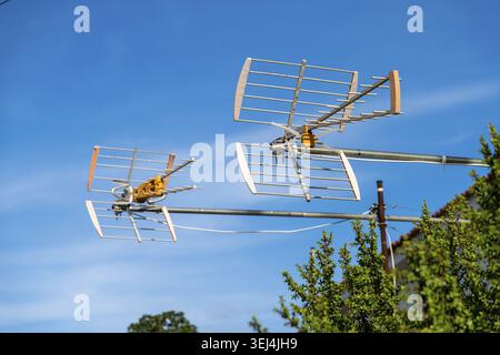 Two metallic TV antennas with yellow connectors extend from a metal pole against a bright blue sky, with green foliage and a tiled roof in the backgro Stock Photo
