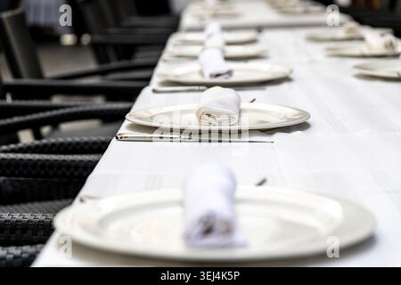 On a table covered with a white tablecloth, white plates, cutlery, and a rolled napkin are stylishly arranged for an elegant table setting Stock Photo