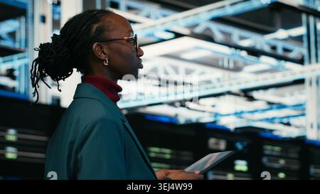 Data center programmer writing code for resource optimization using tablet application. African american woman walking in server hub using programming script on device to upgrade equipment, camera B Stock Photo