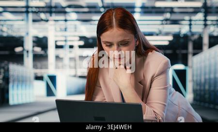 Using laptop, equipment and female programmer at server room workplace desk enhancing operational effectiveness. Monitoring rackmounts, notebook and system administrator in data center, camera B Stock Photo