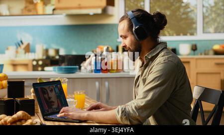 Graphic designer editing photos and wearing headphones at home, seated at kitchen table. Laptop interface shows photo editing software with color grading and retouching tools, visual content, Stock Photo