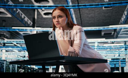 Female programmer at server room workplace desk using laptop to enhance operational effectiveness of equipment. System administrator in data center using notebook to monitor rackmounts, camera A Stock Photo