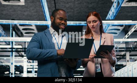 Server room teamworking colleagues using tablet and laptop to enhance operational effectiveness. Engineering team brainstorming in data center, using devices to monitor gear rackmounts, camera B Stock Photo