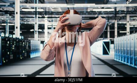 Female technician in server room putting VR goggles on to enhance hardware operational effectiveness. Data center IT expert using virtual reality technology to monitor gear mainframes, camera A Stock Photo
