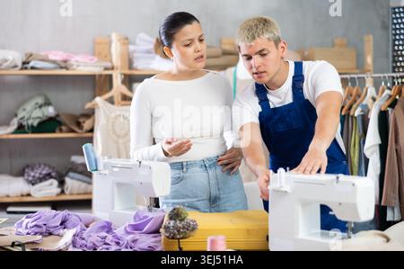 Focused young repairman repairing sewing-machine and Asian tailoress talking Stock Photo