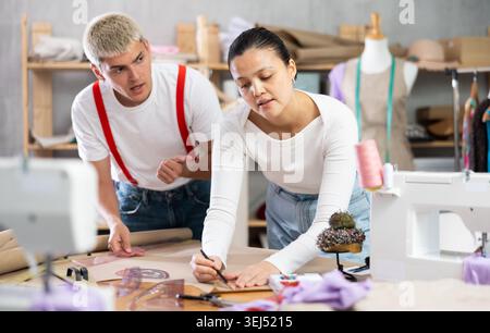 Two seamstresses male and female working in sewing workshop, woman drawing pattern on sheet of paper using curves and man consulting and helping Stock Photo