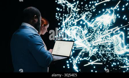 Team of AI startup data scientists implement deep learning algorithms with laptop, collaborating. Coworkers coding, modifying artificial intelligence neural network architecture with device, camera B Stock Photo