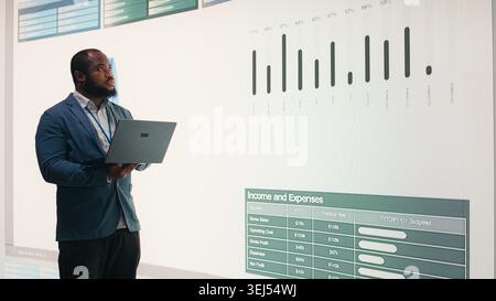 Manager analyzes revenue, expenses and net profit displayed on presentation wall using laptop. African american man uses notebook to review quarterly KPIs results, camera A Stock Photo