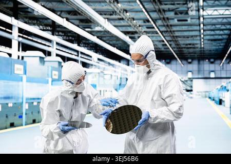 Processing silicon wafer, automation and multiethnic men in automated manufacturing plant. Inspecting semiconductor substrates hardware, bunny suits and coworkers doing quality control Stock Photo