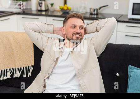 Young man at home leans back and places hands behind head and relaxes muscles with slow breath. Caucasian guy on sofa stretches elbows widens chest and shows calm relief smile and steady mood again Stock Photo