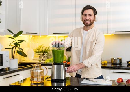 Middle-aged man making banana smoothie in kitchen using blender while listening music for healthy nutrition. Young guy training for fitness mixing fru Stock Photo