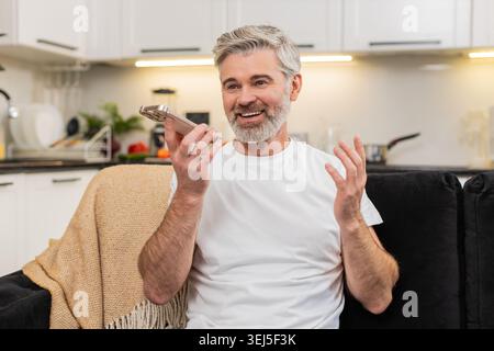 Middle-aged man at home records voice note on smartphone waves hello on speaker and shares news. Mature guy on sofa smiles talks sends chat reply then listens to playback nods and adds a short update. Stock Photo