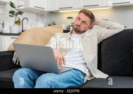 Young man at home works on laptop feels tired and demotivated and takes deep sigh during hard task. Caucasian guy on sofa slumps shoulders shows fatigue stress low energy and tries to focus now again Stock Photo
