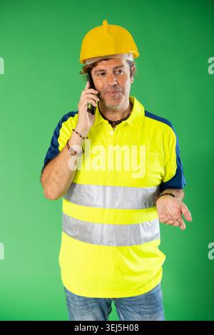 Adult man in reflective polo shirt and hard hat talking on mobile phone on green screen Stock Photo