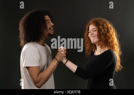Diverse couple holding hands and laughing on a dark background, symbolizing togetherness Stock Photo