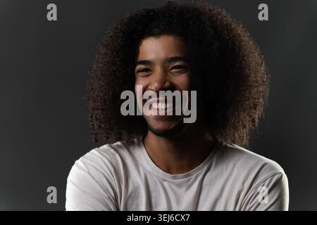 Young man with afro hair joyfully smiling against a dark background, reflecting happiness Stock Photo