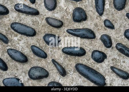 Black pebbles rest on rough concrete surface. They vary in size and shape. Pebbles appear wet and smooth. Texture contrasts between stone and concrete Stock Photo