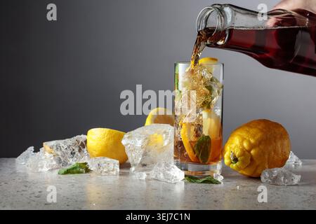 Iced black tea is poured from a bottle into a tall glass with lemon wedges, ice cubes and mint leaves. Gray gradient background with copy space. Stock Photo