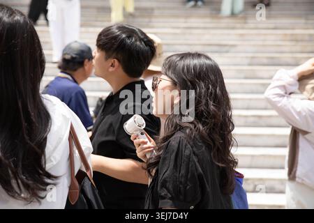 BANGKOK, THAILAND-MAY 16, 2026: Female tourist using handheld electric fan blowing face, casual travel walking through foreign city, side profile. Stock Photo