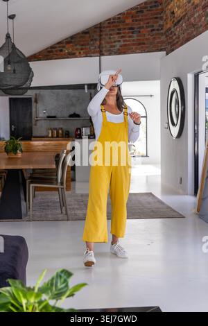 Woman wearing white VR headset and yellow overalls stepping, reaching toward dining table at home Stock Photo
