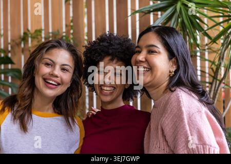 Diverse female friends posing on patio, leaning against wooden slat fence with potted plants Stock Photo