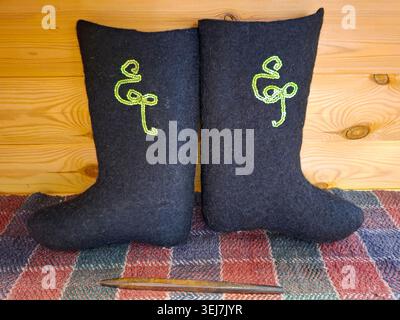 A pair of traditional Russian black felt boots sits on a woven rug. The boots are decorated with green sequins. The background shows warm wooden walls Stock Photo
