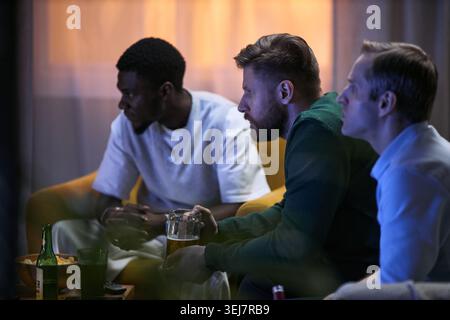 Multiethnic young adult men watching television together, sharing leisure time and male friendship at home. Useful for campaigns about social bonding, lifestyle marketing, medium shot Stock Photo
