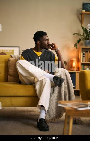 Vertical shot of young Black man sitting thoughtfully on sofa, looking away and reflecting on friendship and support. Ideal for mental wellbeing, counseling, lifestyle campaigns Stock Photo