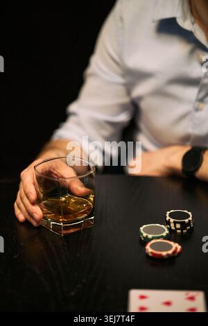 Vertical shot of young adult man holding whiskey glass beside poker chips and playing card, suggesting gambling habit and risk. Suitable for addiction awareness, betting, debt, mens lifestyle themes Stock Photo