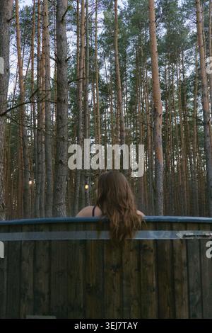 Back view of a beautiful woman with long, dark hair sitting in the outdoor, wood-fired jacuzzi. Coniferous forest in the background. Stock Photo