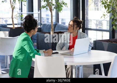 Diverse female coworkers sitting at round table in lounge, green blazer, using laptop and phone Stock Photo