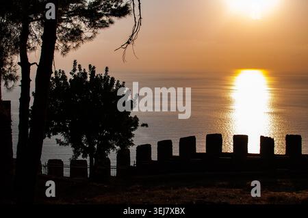 A dramatic silhouette of the ancient fortress wall and pine trees against a bright golden sunset over the calm Mediterranean Sea in Alanya, Turkey. Stock Photo