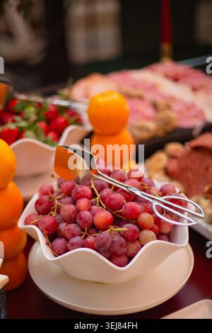 Bowl of red grapes with serving tongs, surrounded by stacked oranges, strawberries, cheese, and charcuterie on a banquet table. Stock Photo