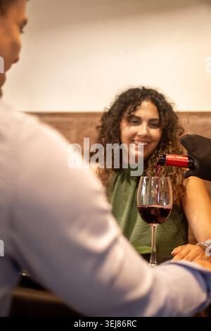 Waiter serving red wine to a happy young couple during a romantic dinner at a cozy restaurant. Warm ambient lighting, elegant table setting and authen Stock Photo