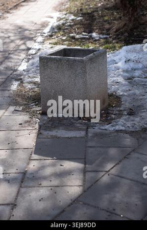 empty square concrete planter sitting on a gray urban sidewalk beside melting snow and scattered litter, casting angular shadows across paving stones; Stock Photo