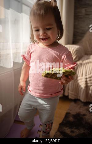 smiling toddler girl in a pink shirt holding a green textured snack by a sunlit window in a cozy living room, warm beige tones, soft natural light, ca Stock Photo
