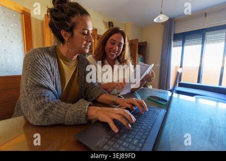 Two women collaborating at home, with one typing on a laptop while the other gestures towards financial documents, brainstorming ideas Stock Photo